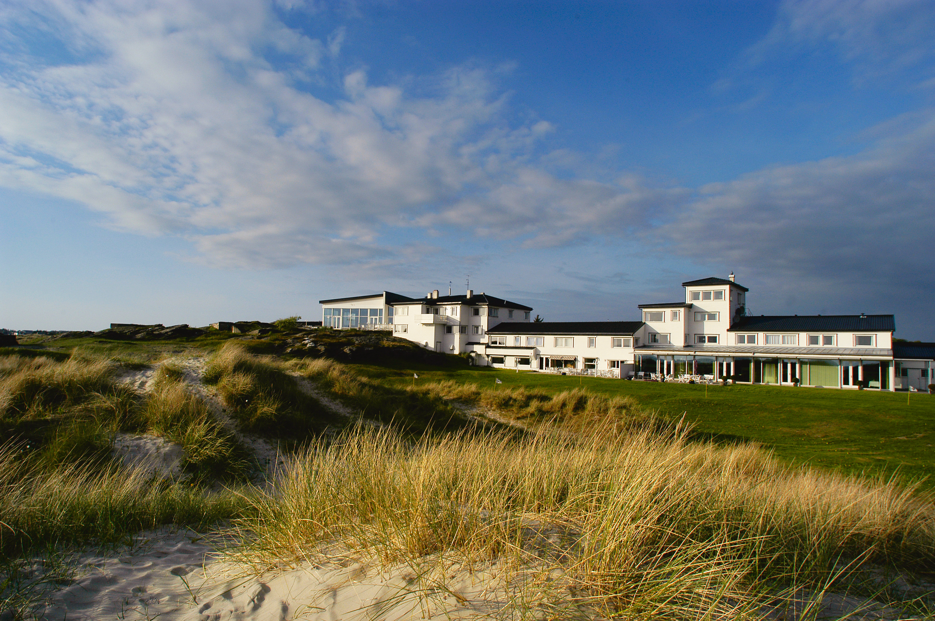 en hvit bygning i bakgrunnen, blå og vid himmel, i forgrunnen sandstrand og strå