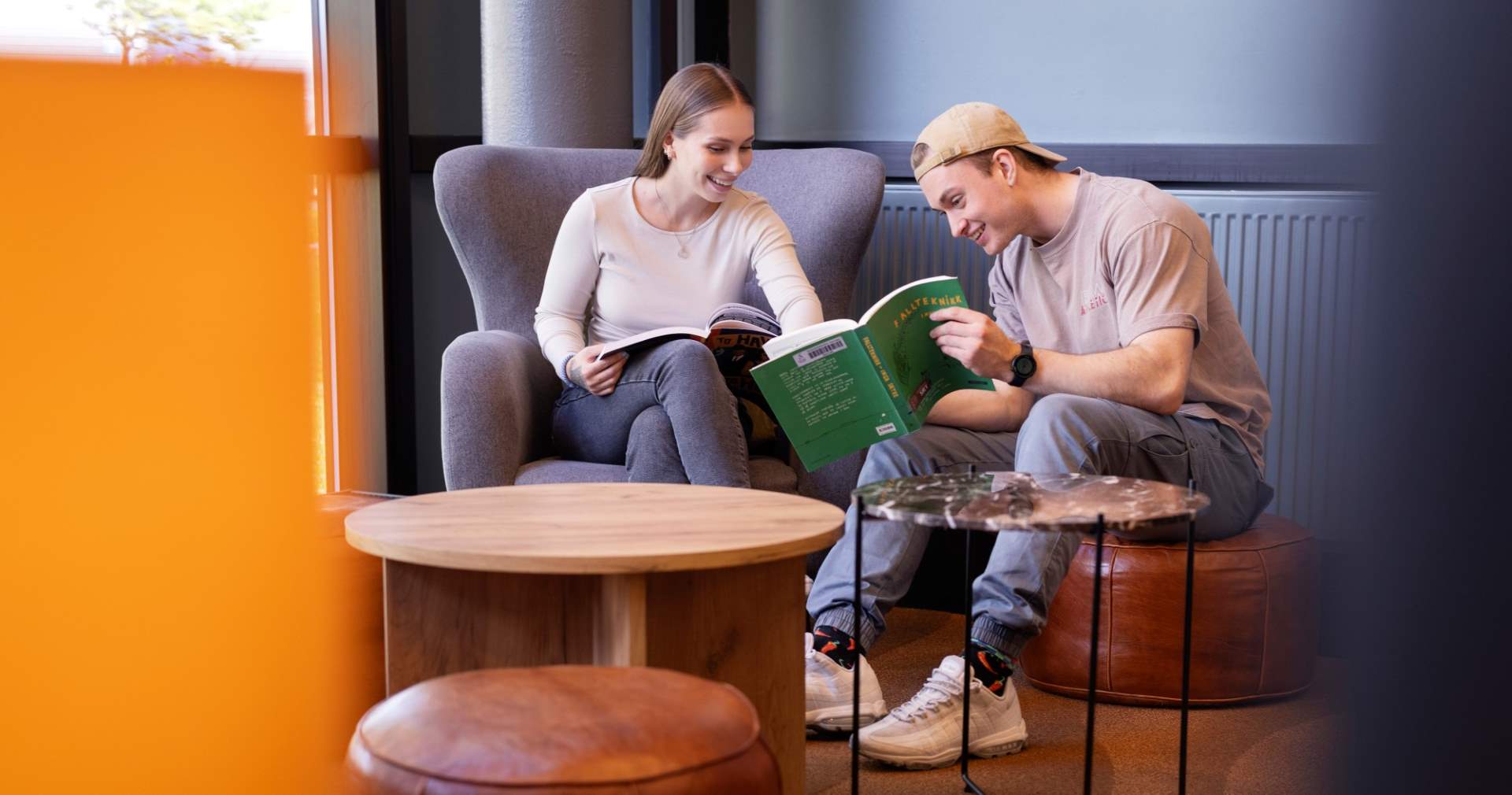 To smilende studenter sitter i stoler ved siden av hverandre og kikker i en bok.