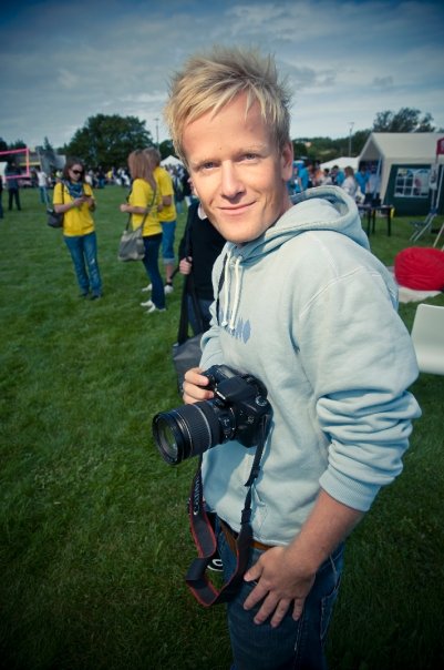 Fotografi av blond mann i lys hettegenser med et kamera i hendene. I bakgrunnen ses  mennesker på en gressplen.