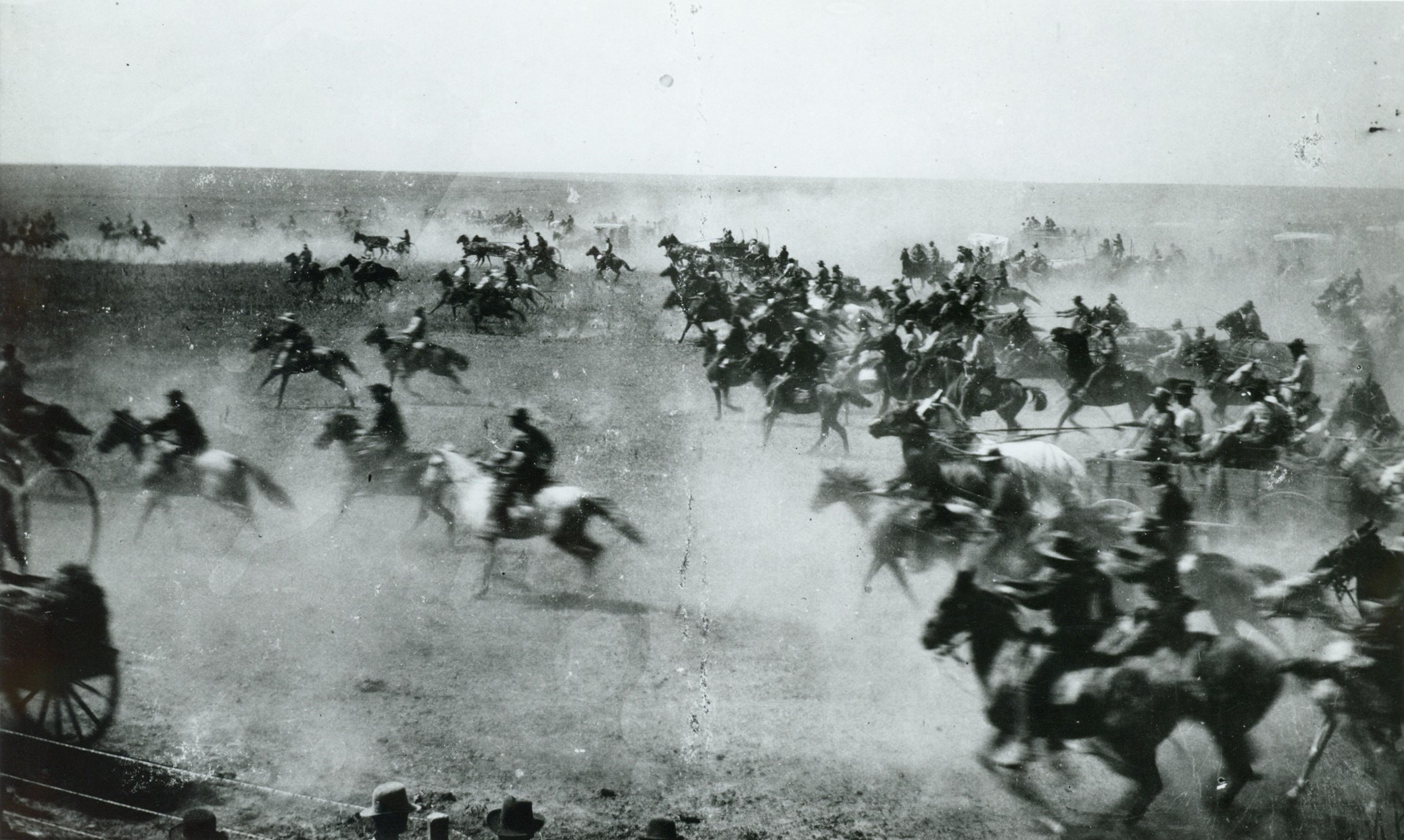 Horses and riders, and horse-drawn wagons kick up dust as they gallop across a flat plain