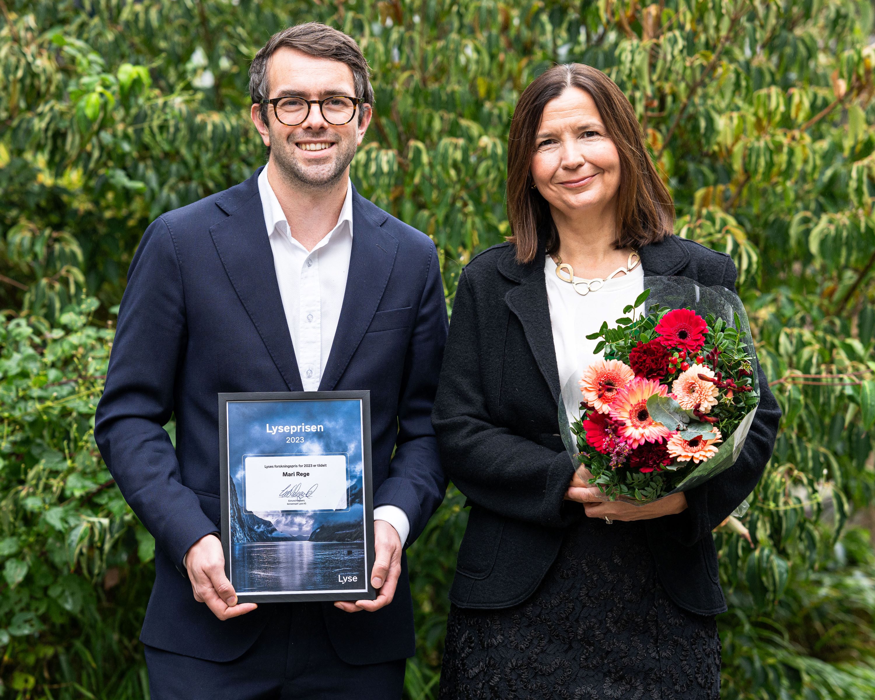 Kvinne og mann foran grønne busker holder et diplom og blomsterbukett i hendene.