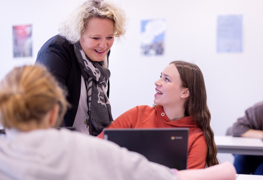 Lærer og elev i munter samtale over en pc. En annen elev sitter med ryggen til og konsentrerer seg.