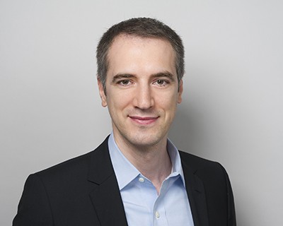 Portrait of a smiling man in a black suit and blue shirt in front of a white wall