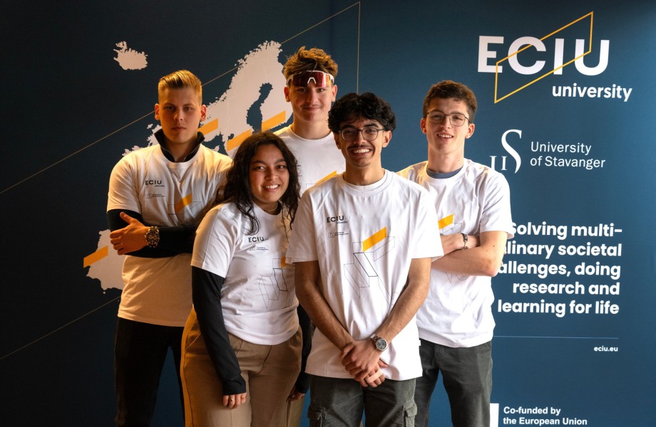 Fem unge studenter i hvite t-skjorter oppstilt foran en blå vegg med skrift og kart over Europa.