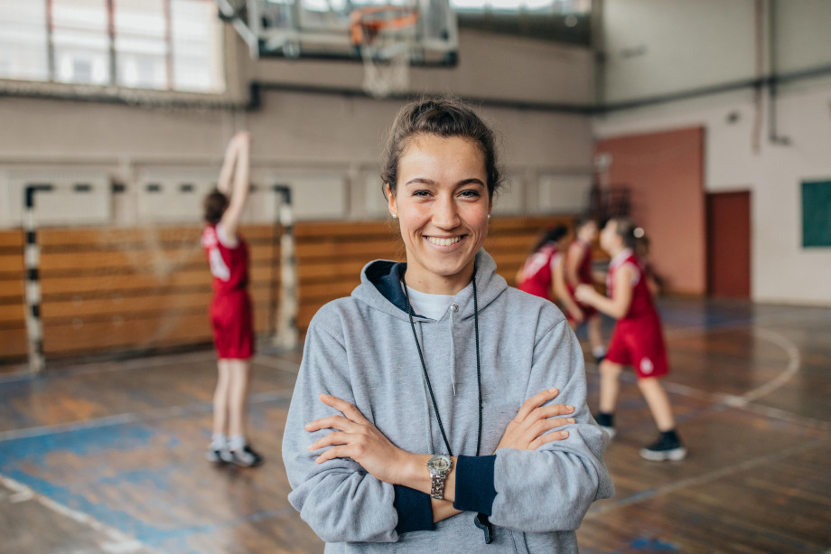 ung kvinne med med håret i hestehale står i en gymsal, smiler til kamera. Tre barn holder på med ball i bakgrunnen