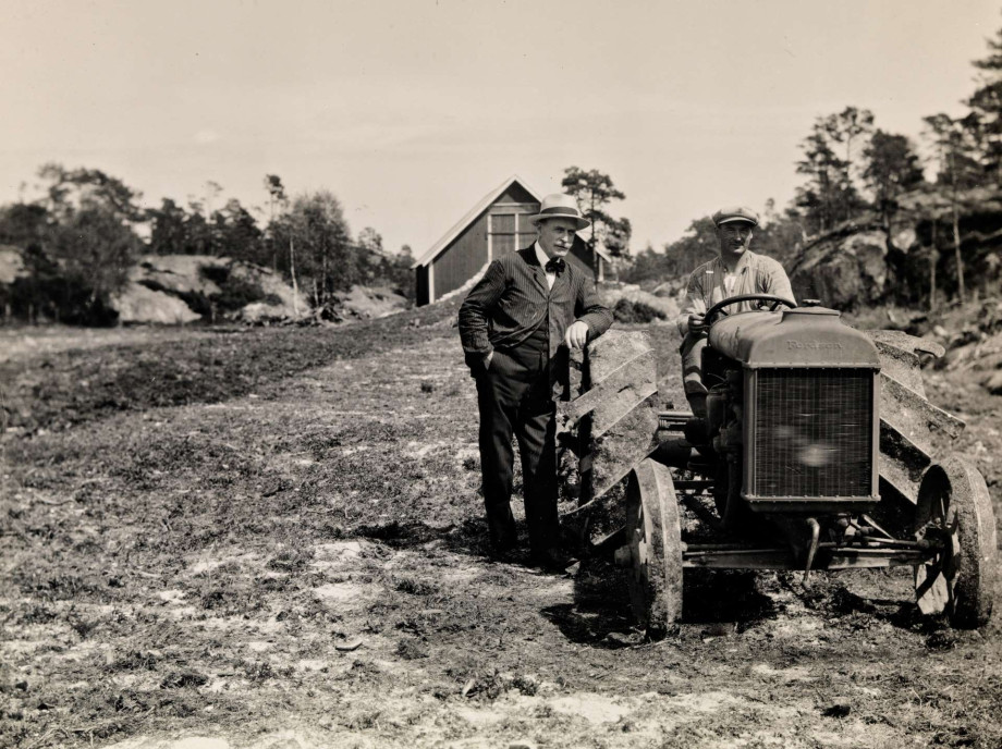 A man in a suit and hat is standing next to a tractor with it's driver. Photo taken in the late 1920s.