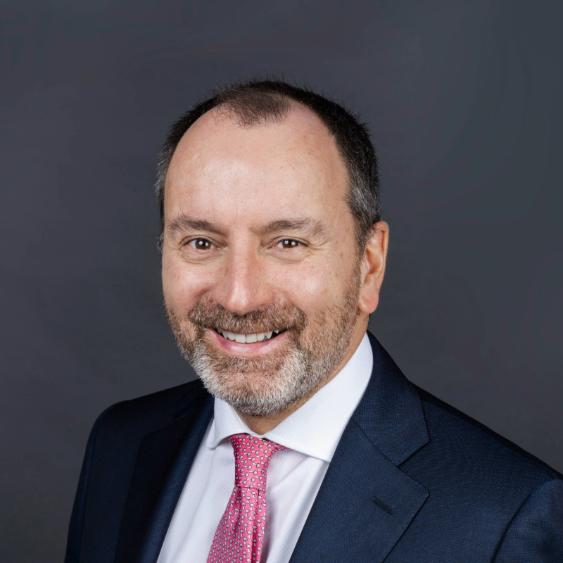 Man in a suit smiles to the camera. Dark background.