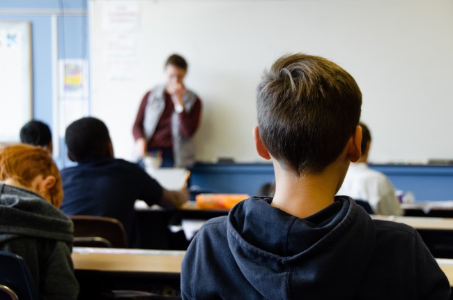 Bakhodet til en gutt. Lenger framme ser vi flere elever ved skolepulten og en lærer foran en hvit tavle. 