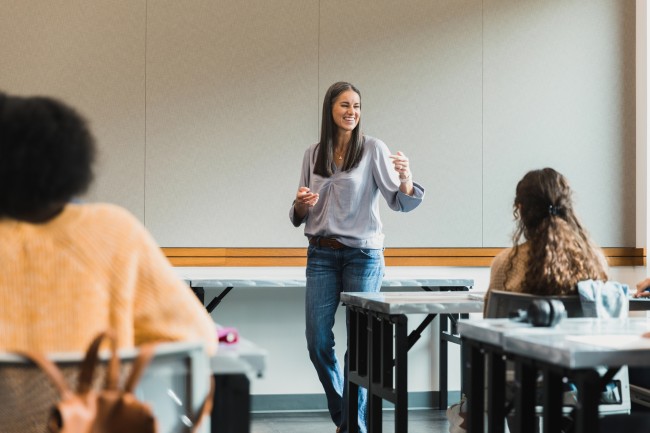 Kvinne står foran gruppe i klasserom. To personer sitter med ryggen til.